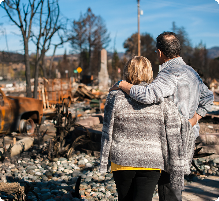 A couple embraces while surveying the aftermath of a fire, surrounded by debris and charred remains of buildings and vehicles.